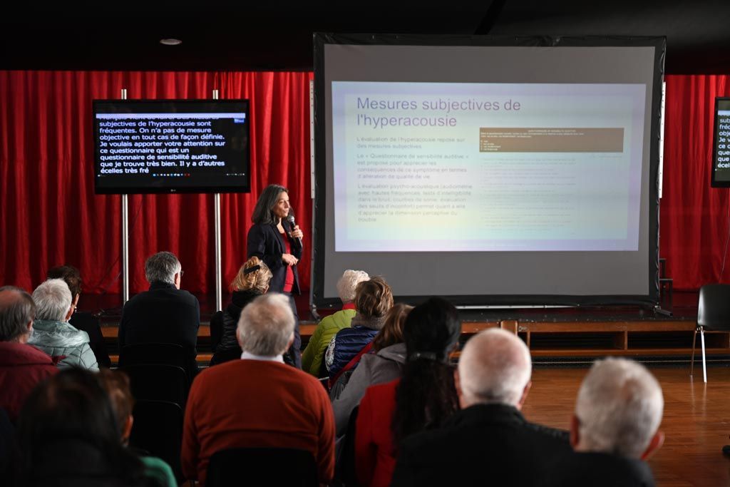 Une femme présente un diaporama dans une salle où se trouve un public. On aperçoit des rideaux rouges et un petit écran.