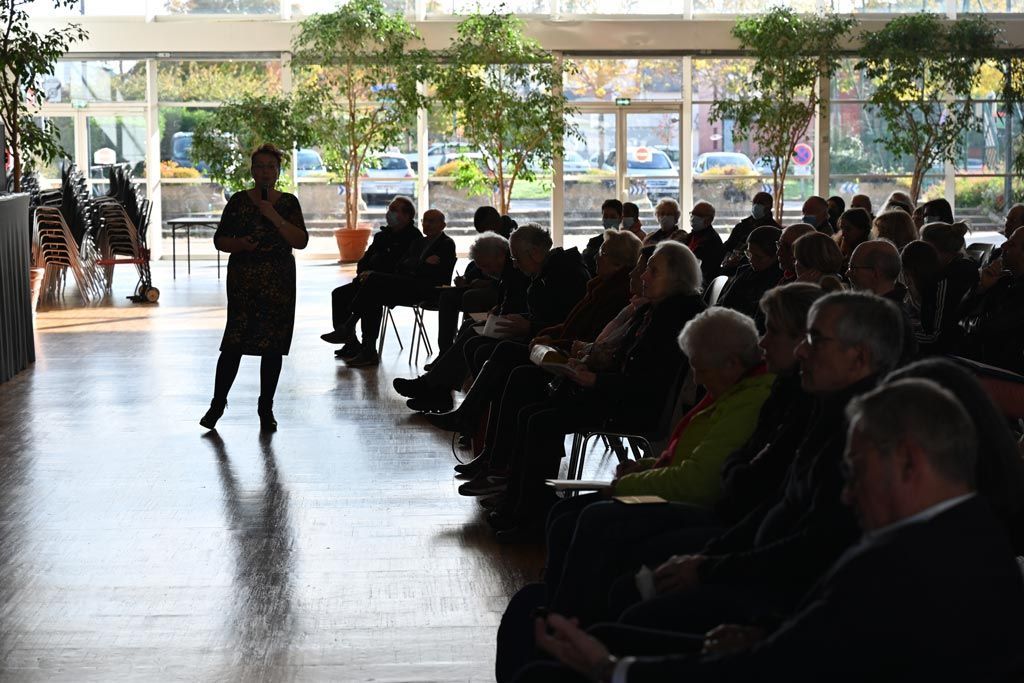 Une femme présente un exposé devant un public assis dans une pièce lumineuse avec de grandes fenêtres et des arbres à l'extérieur.
