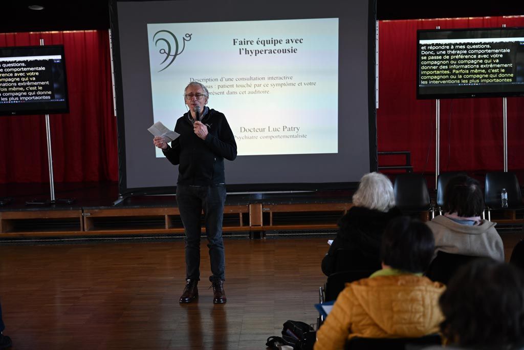 Une personne fait une présentation devant un public assis. Des écrans affichent du texte.