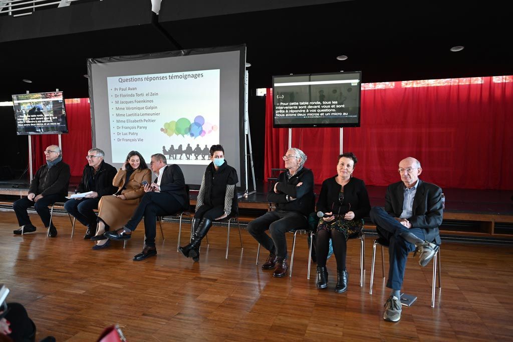 Un panel de huit personnes assises sur scène, présentant un exposé au public, avec un écran de projection derrière elles.