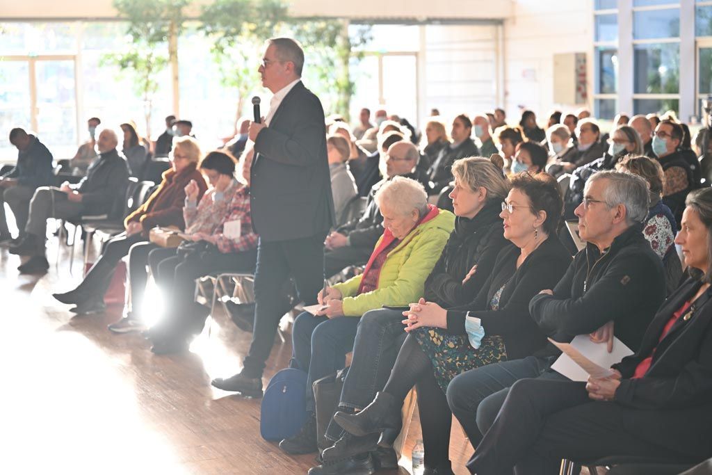 Un homme s'adresse à un public assis dans une salle lumineuse. Beaucoup de personnes portent des masques.