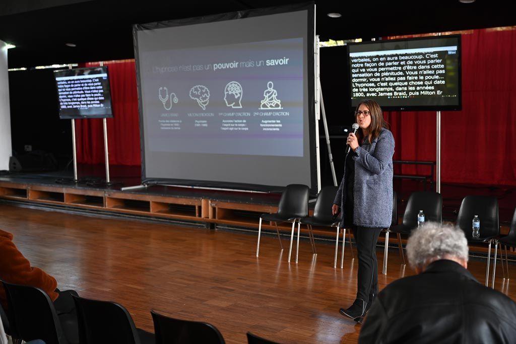 Femme présentant un exposé depuis un podium, avec un écran de projection. Intérieur décoré de rideaux rouges et de chaises vides.