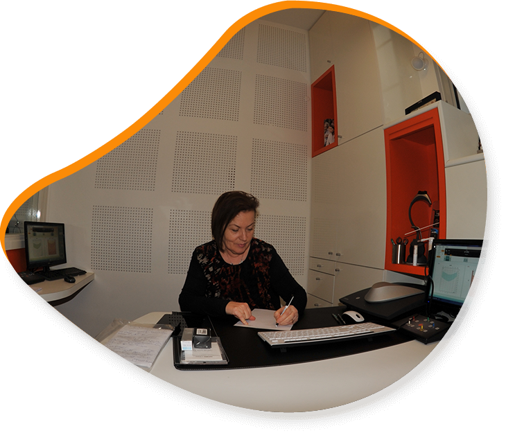 Une femme écrit à un bureau dans un bureau aux accents orange. Murs blancs avec panneaux acoustiques. Ordinateur sur le bureau.