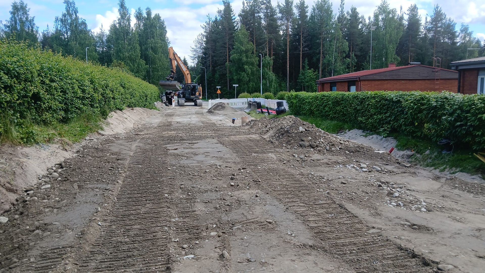 Rakenteilla oleva hiekkatie, kaivinkone, työntekijöitä ja pensasaitoja metsäalueen vieressä