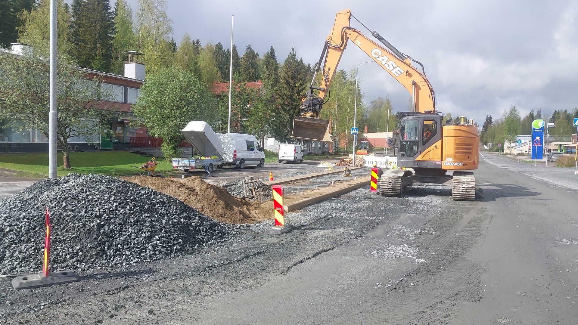 Tienrakennuskohta kaivinkoneen, sorakasojen ja liikennekartioiden kanssa tien laidassa pilvisen taivaan alla