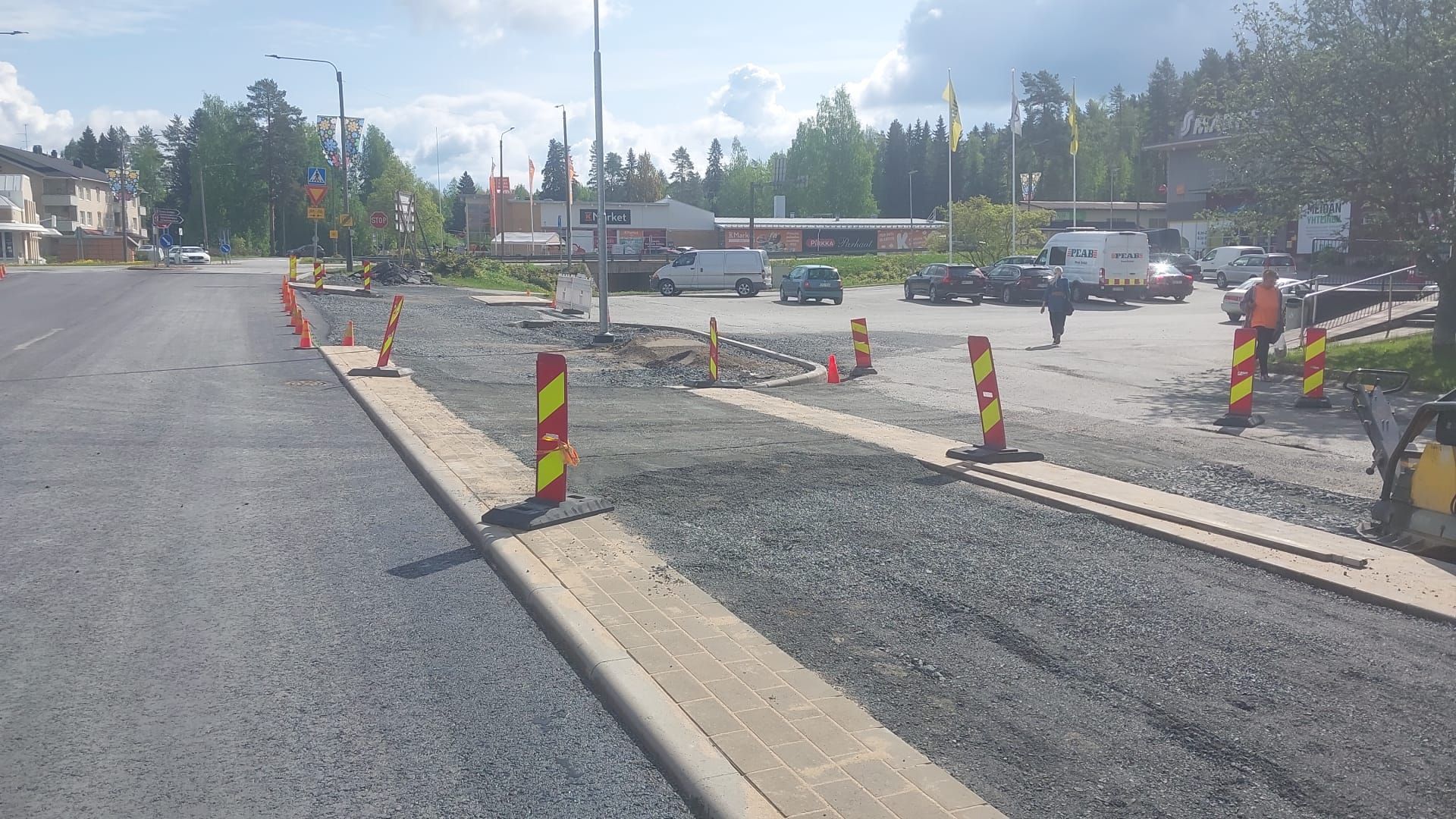 Tienrakennustyömaa, jossa on tuoretta asfalttia, kartioita ja raidallisia barrikadeja pysäköityjen autojen vieressä pilvisen taivaan alla