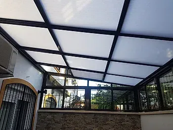 Patio cerrado con techo de vidrio, marco negro y paneles que filtran la luz.
