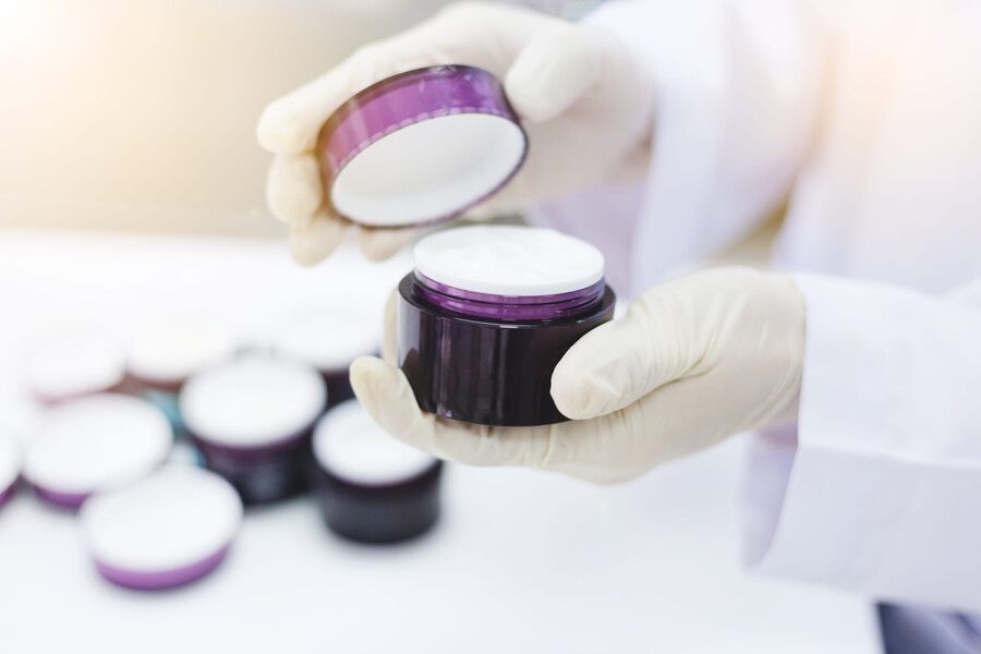 Una persona con bata blanca de laboratorio y guantes sostiene un frasco de cosméticos morado abierto, lleno de crema blanca.