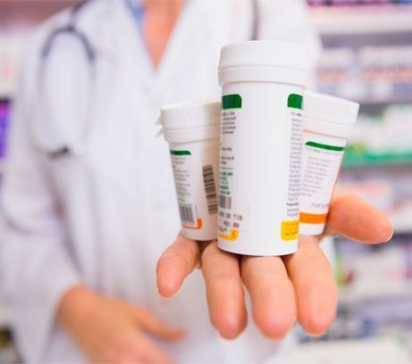 Una persona con bata blanca de laboratorio sostiene tres frascos de medicamentos recetados en su mano abierta frente a una farmacia borrosa.