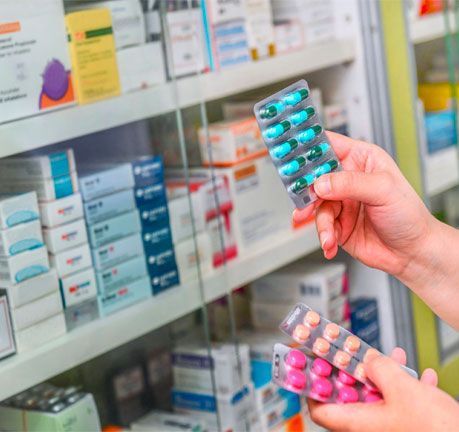 En una farmacia, una persona sostiene blísteres de cápsulas y comprimidos azules, rosas y beige frente a los estantes de medicamentos.