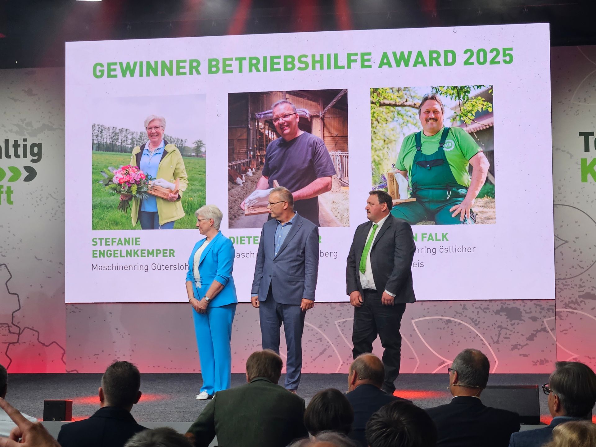 Preisverleihung: Drei Personen auf der Bühne, dahinter Gewinnerfotos. Publikum im Vordergrund.