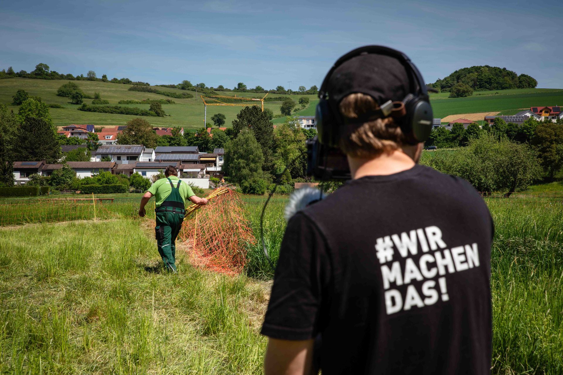 Eine Person mit Kopfhörern auf dem Rücken beobachtet eine andere Person, die orangefarbenes Heu über eine Wiese trägt, im Hintergrund ist eine Stadt zu sehen.