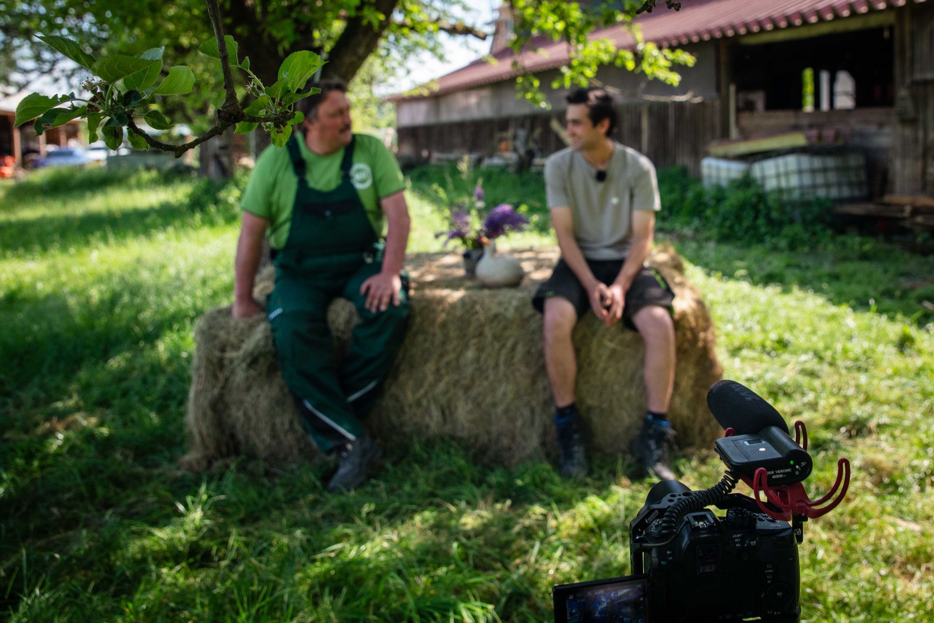 Zwei Männer sitzen während eines Interviews im Freien auf einem Heuballen, im Vordergrund ist eine Kamera zu sehen. Ein Mann trägt einen grünen Overall, der andere ein T-Shirt.