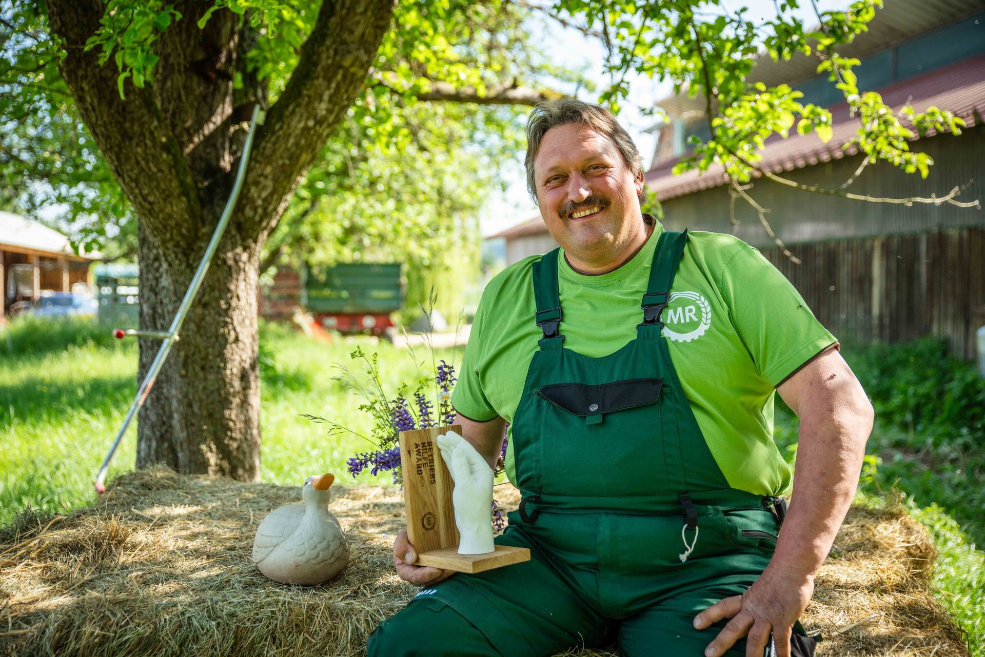 Lächelnder Bauer sitzt auf Heuballen und hält einen Holzblock mit Blumen. Er trägt einen grünen Overall im Freien.