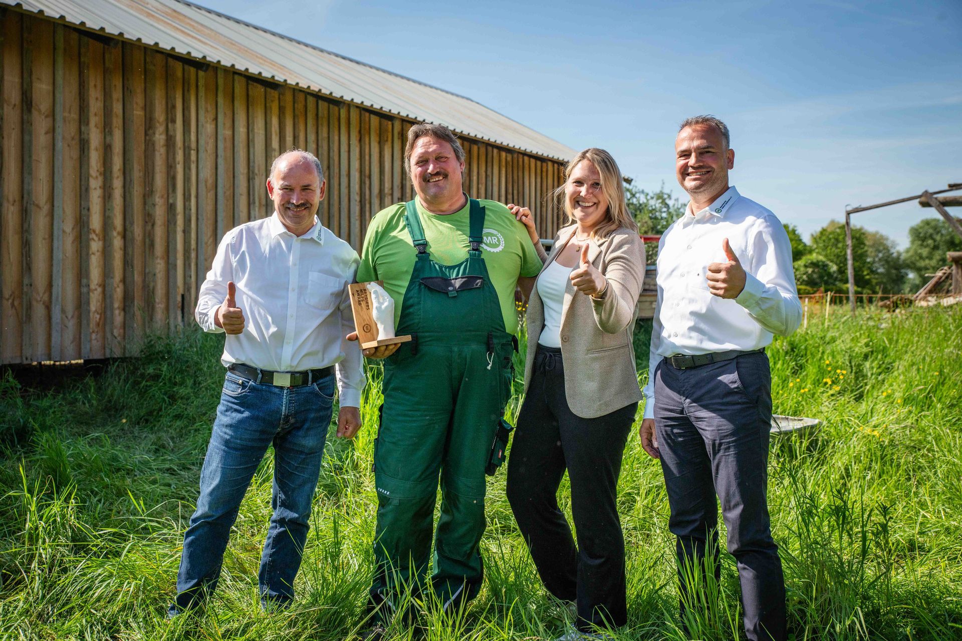 Vier Personen stehen draußen vor einem Holzgebäude und strecken alle den Daumen nach oben. Sie befinden sich an einem sonnigen Tag auf einer Rasenfläche.