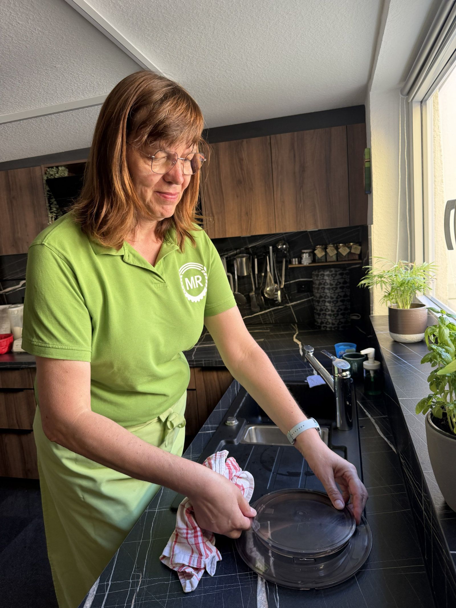 Frau in grünem Hemd trocknet eine Pfanne an einem Küchenspülbecken neben einem hellen Fenster.