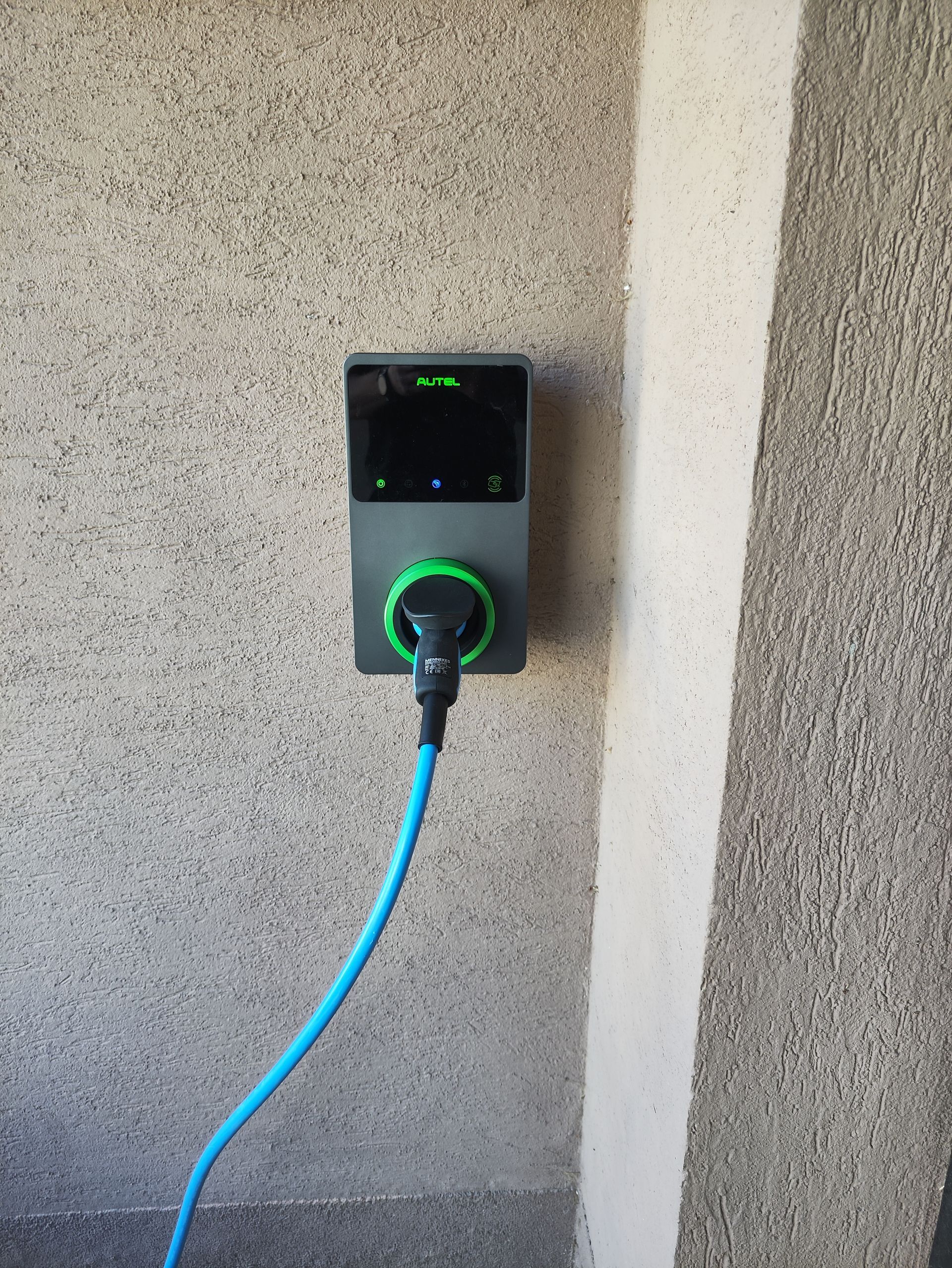 Borne de recharge pour véhicule électrique fixée au mur, avec un câble bleu branché, contre un mur gris texturé.