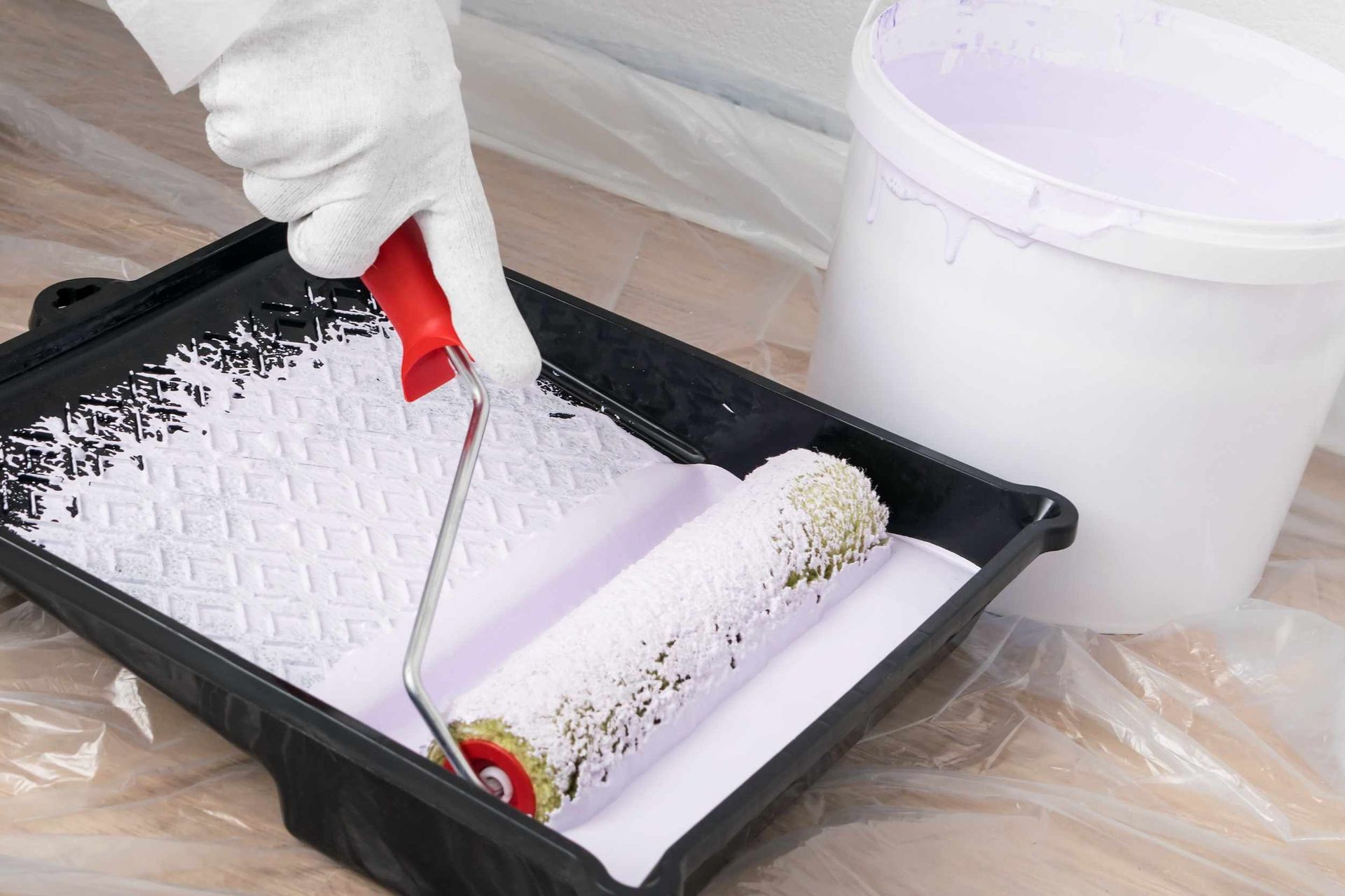 Persona con guantes blancos que utiliza un rodillo de pintura para aplicar pintura violeta claro en una bandeja de pintura, al lado de un balde de pintura.