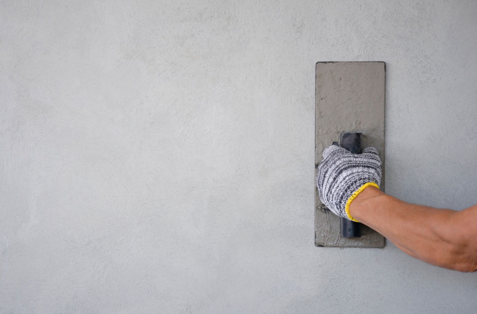 Persona con una mano enguantada que utiliza una paleta para alisar yeso en una pared gris.