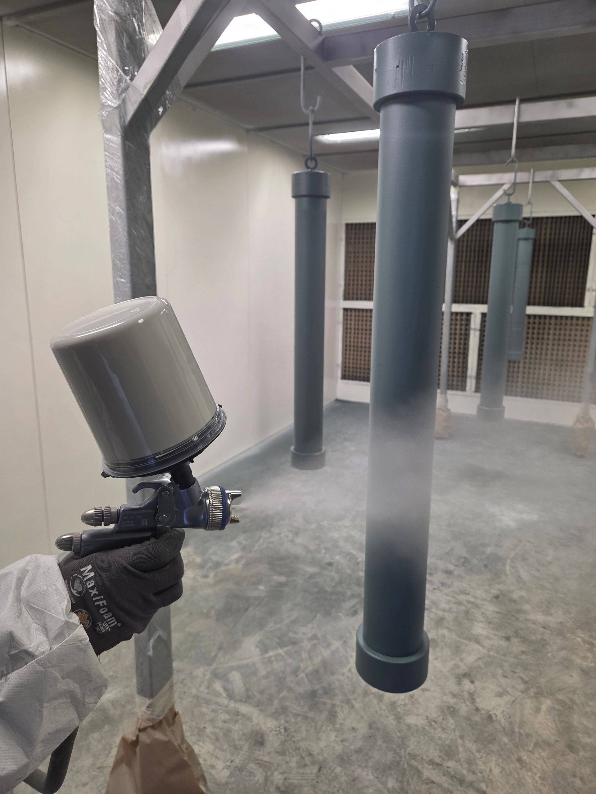 Dans une cabine de peinture industrielle, une main gantée utilise un pistolet à peinture pour recouvrir des tuyaux métalliques cylindriques gris suspendus.