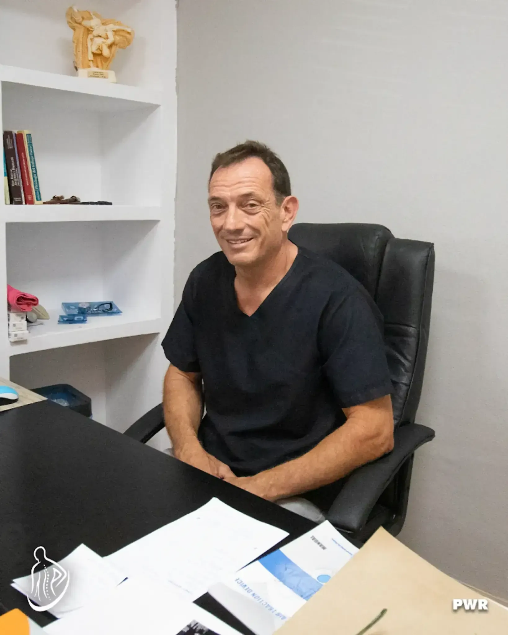 Hombre con camisa negra sentado en un escritorio, sonriendo. Oficina con estantería al fondo.