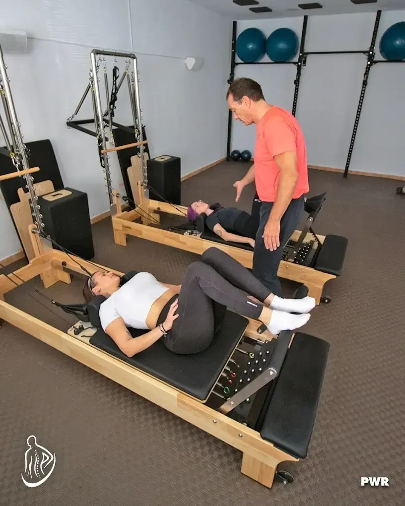 Dos personas realizando ejercicios de Pilates en máquinas, con el instructor guiándolos, en el interior.