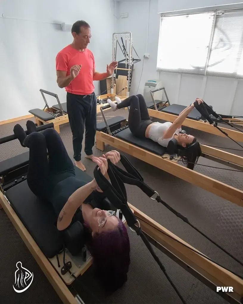 Un instructor de pilates guía a dos personas en reformers. Lleva camisa roja. Estudio con luz natural.
