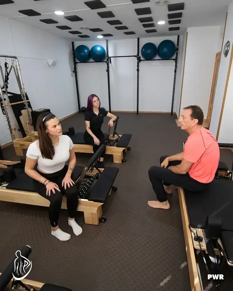 Tres personas en un estudio de pilates, sentadas en máquinas, conversando. Pelotas de ejercicio azules en la pared.