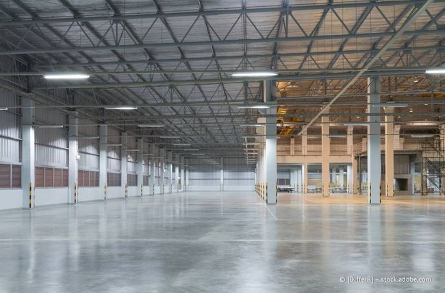 Innenansicht eines großen, leeren Lagerhauses mit Betonboden, Metallträgern und Deckenbeleuchtung.