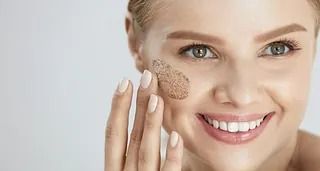Una mujer sonríe mientras se aplica un exfoliante en la cara.