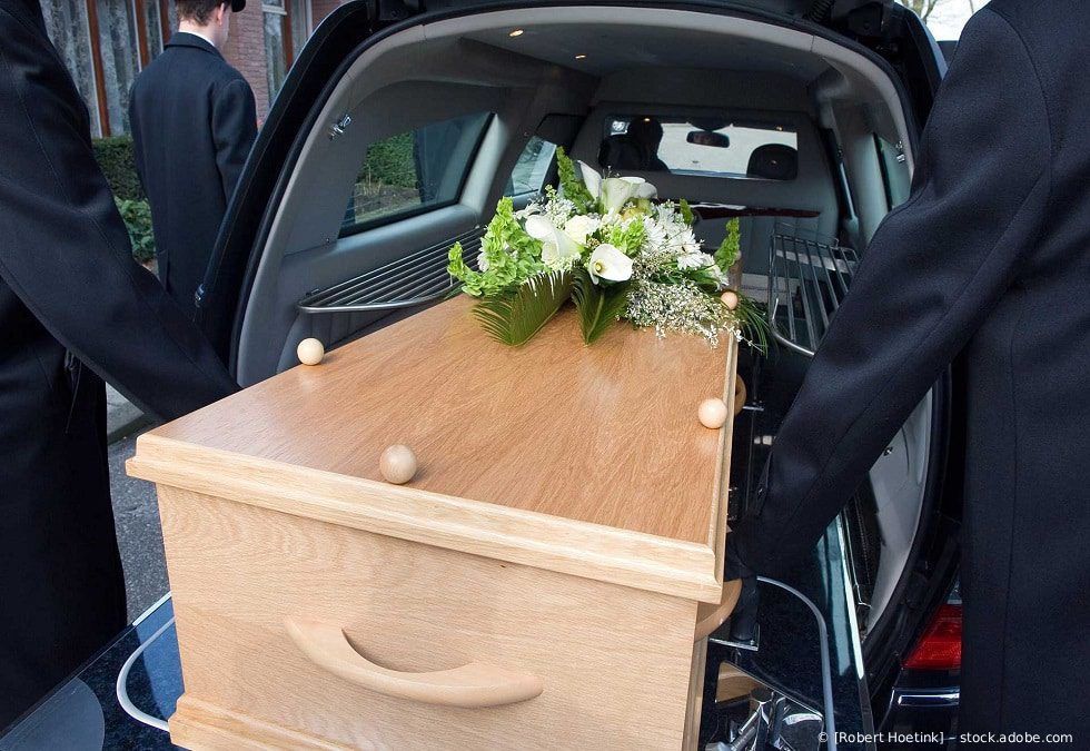 Ein Holzsarg wird in ein Auto verladen; er ist mit weißen Blumen bedeckt und wird von zwei Trauernden gehalten.