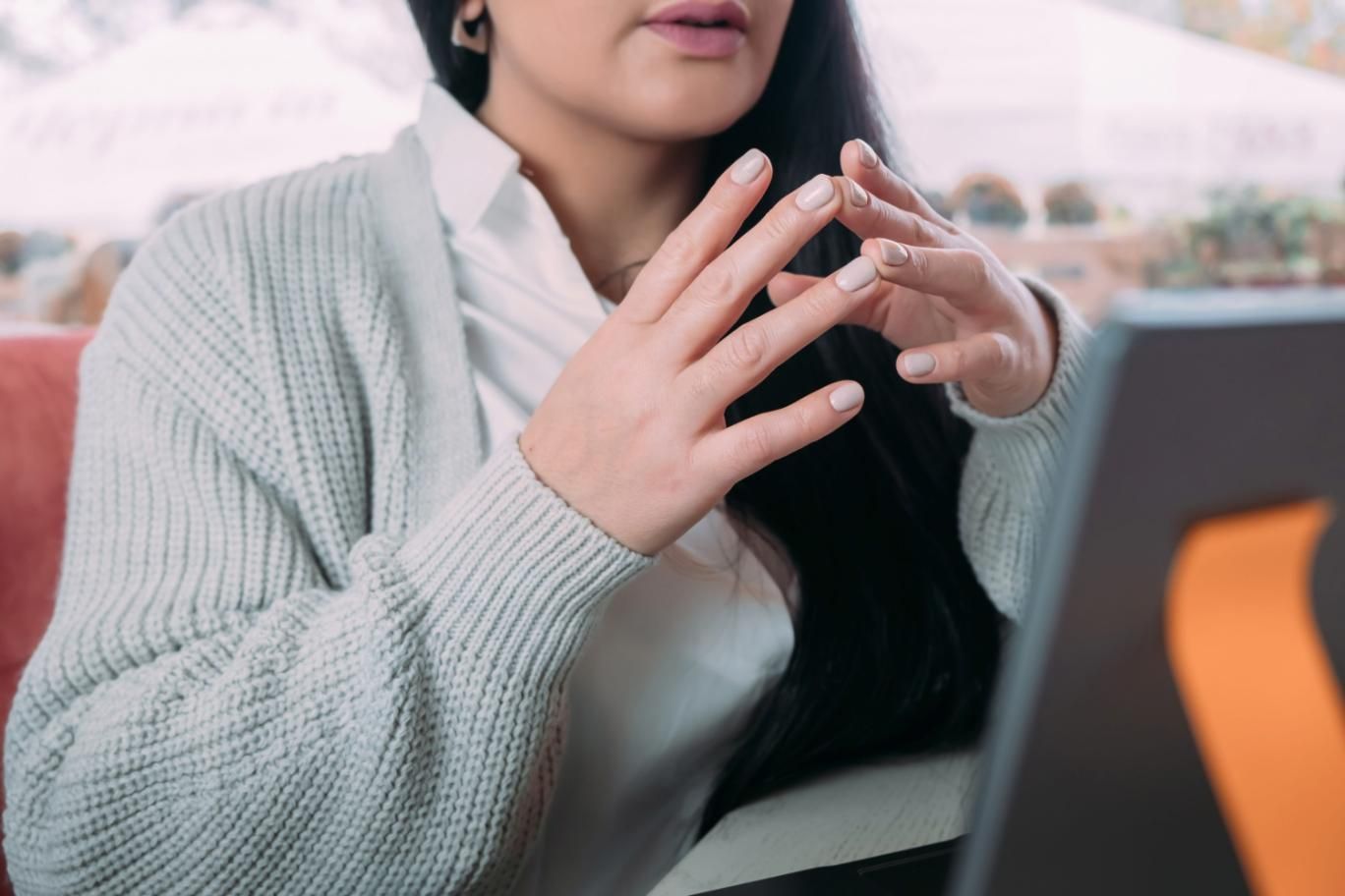 Una persona que lleva un suéter gris claro gesticula con las manos mientras está sentada a una mesa con una computadora