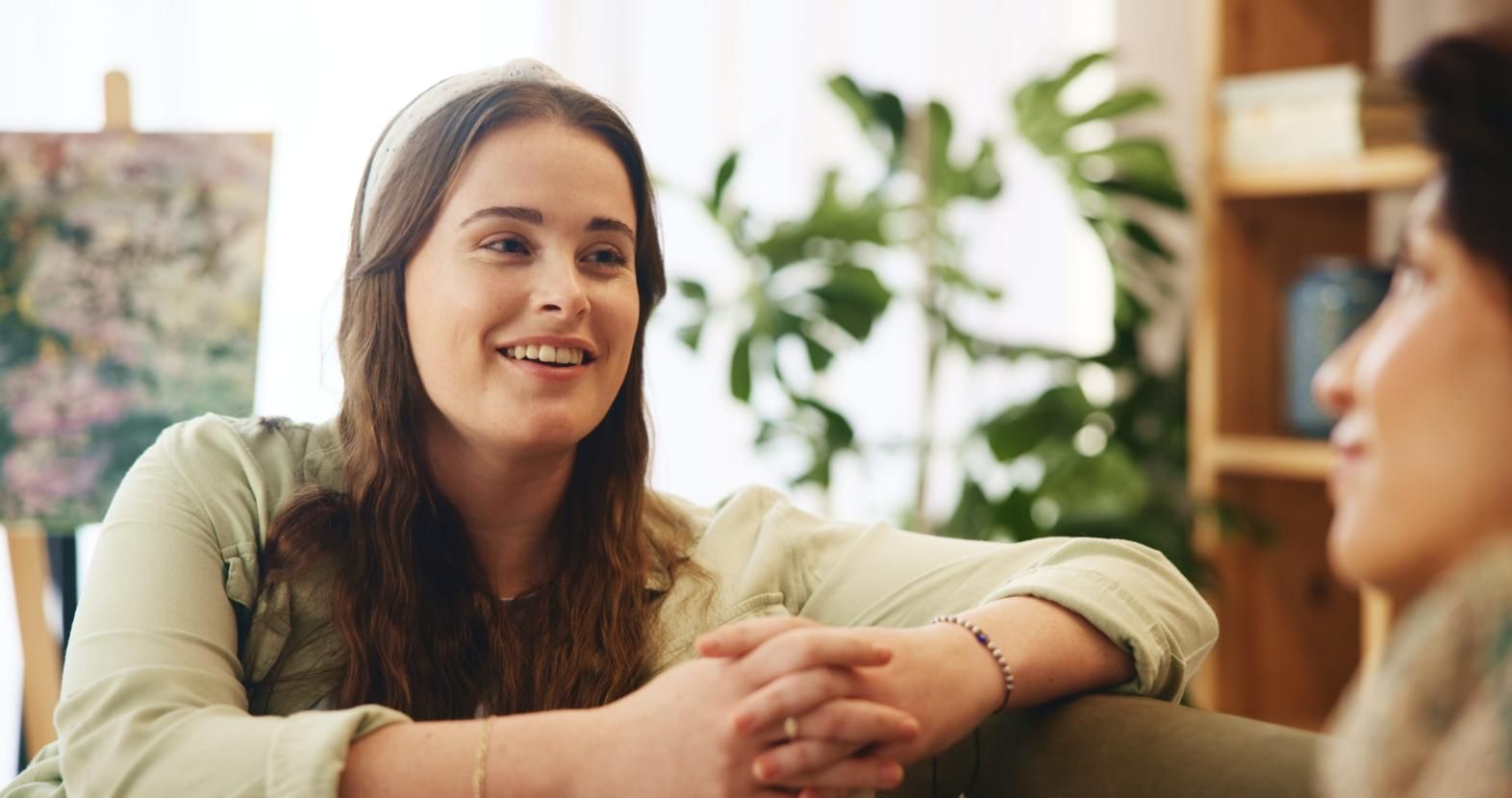 Dos personas sentadas juntas en una habitación luminosa y llena de plantas, una de ellas hablando y sonriendo.