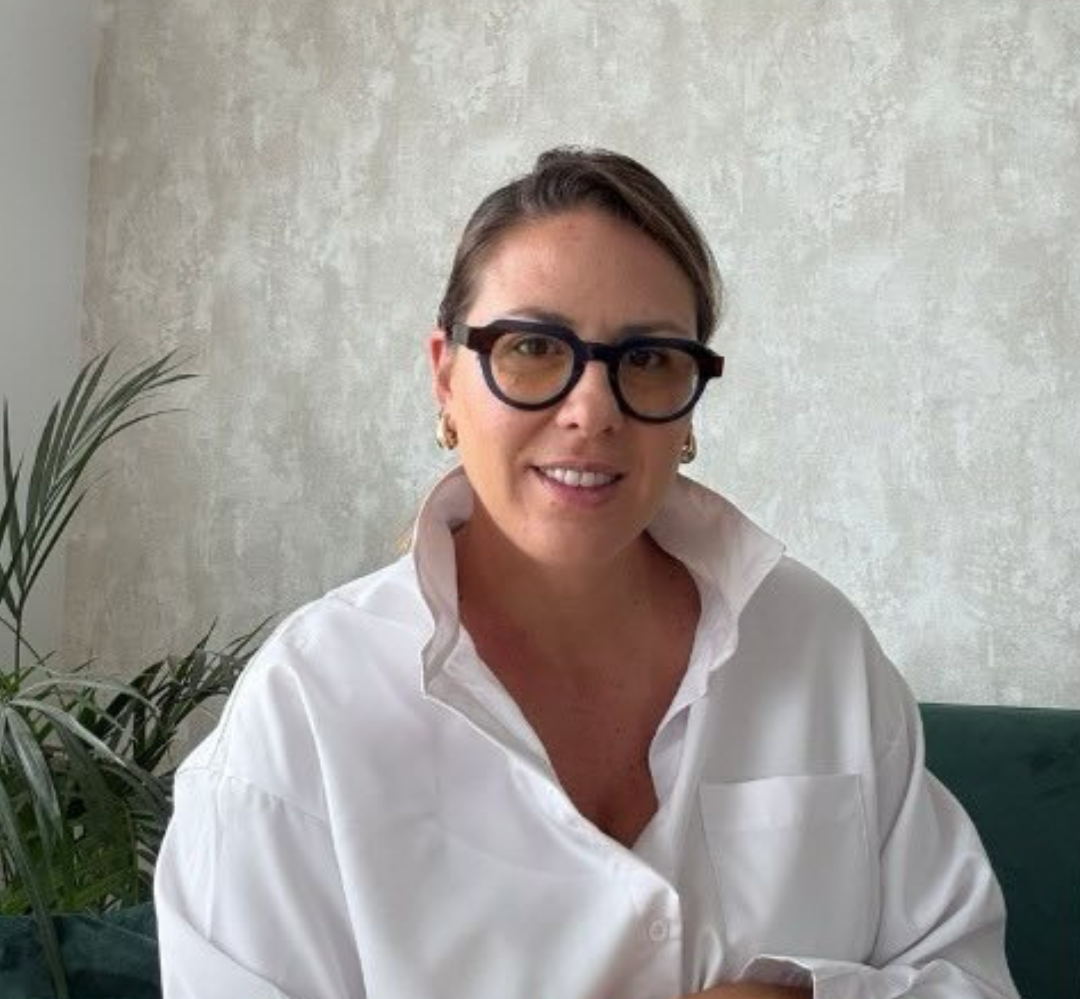 Una persona con camisa blanca de cuello y gafas de montura negra, sonriendo en una habitación con una planta