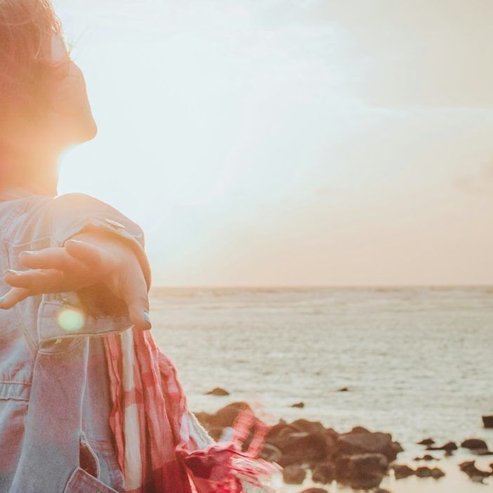 Una persona con los brazos extendidos permanece de pie cerca de una costa rocosa, contemplando 