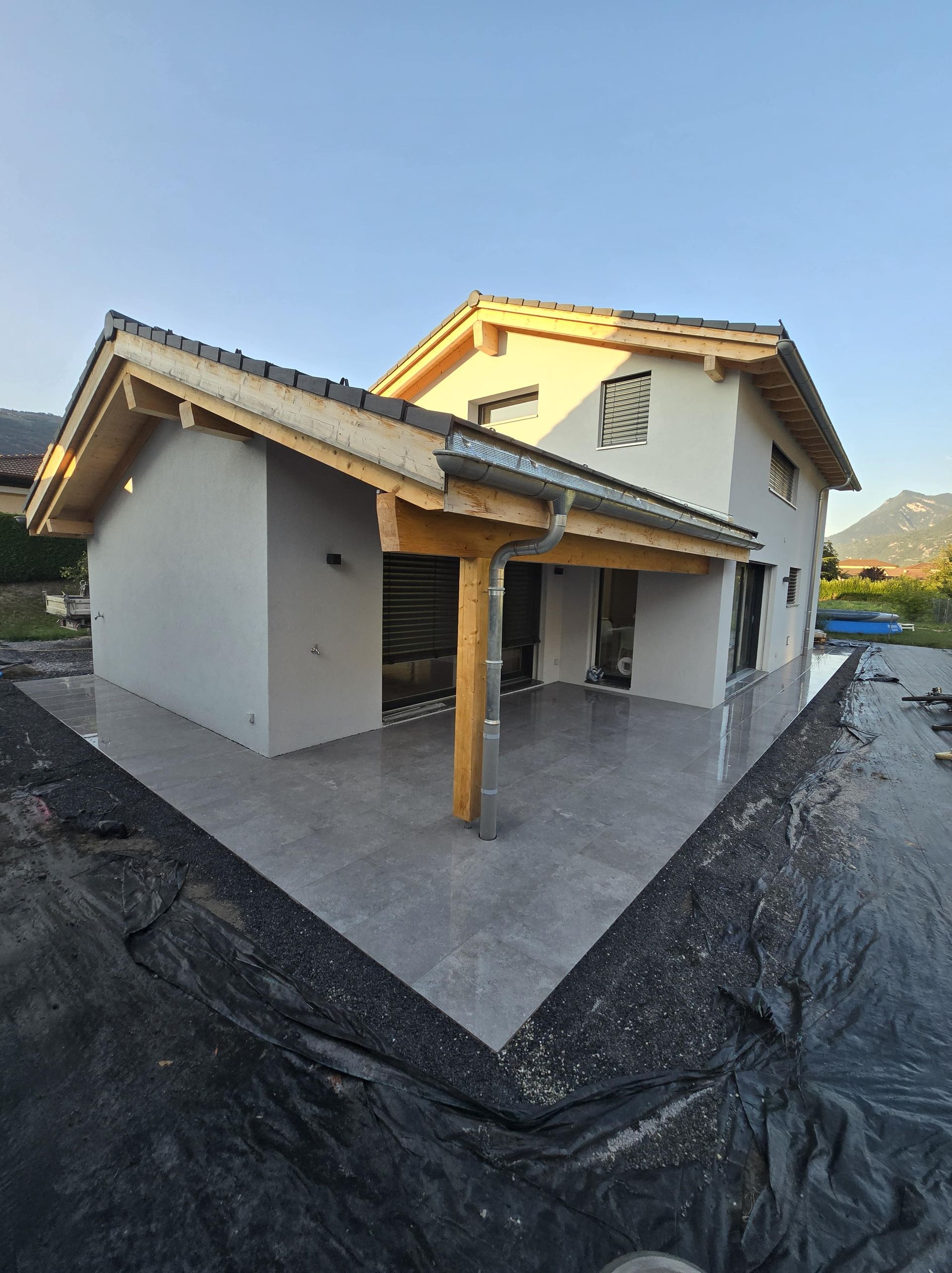 vue extérieure d'une maison en construction avec une terrasse dallée et une bâche sur du gravier autour
