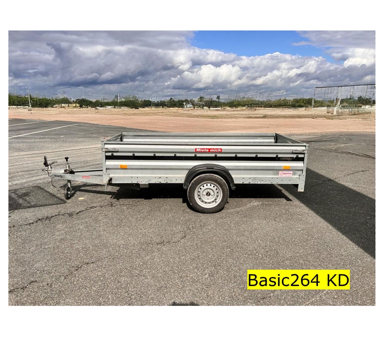 Ein silberner Nutzanhänger auf Asphalt mit einem bewölkten Himmel im Hintergrund. Aufschrift: „Basic264 KD“.