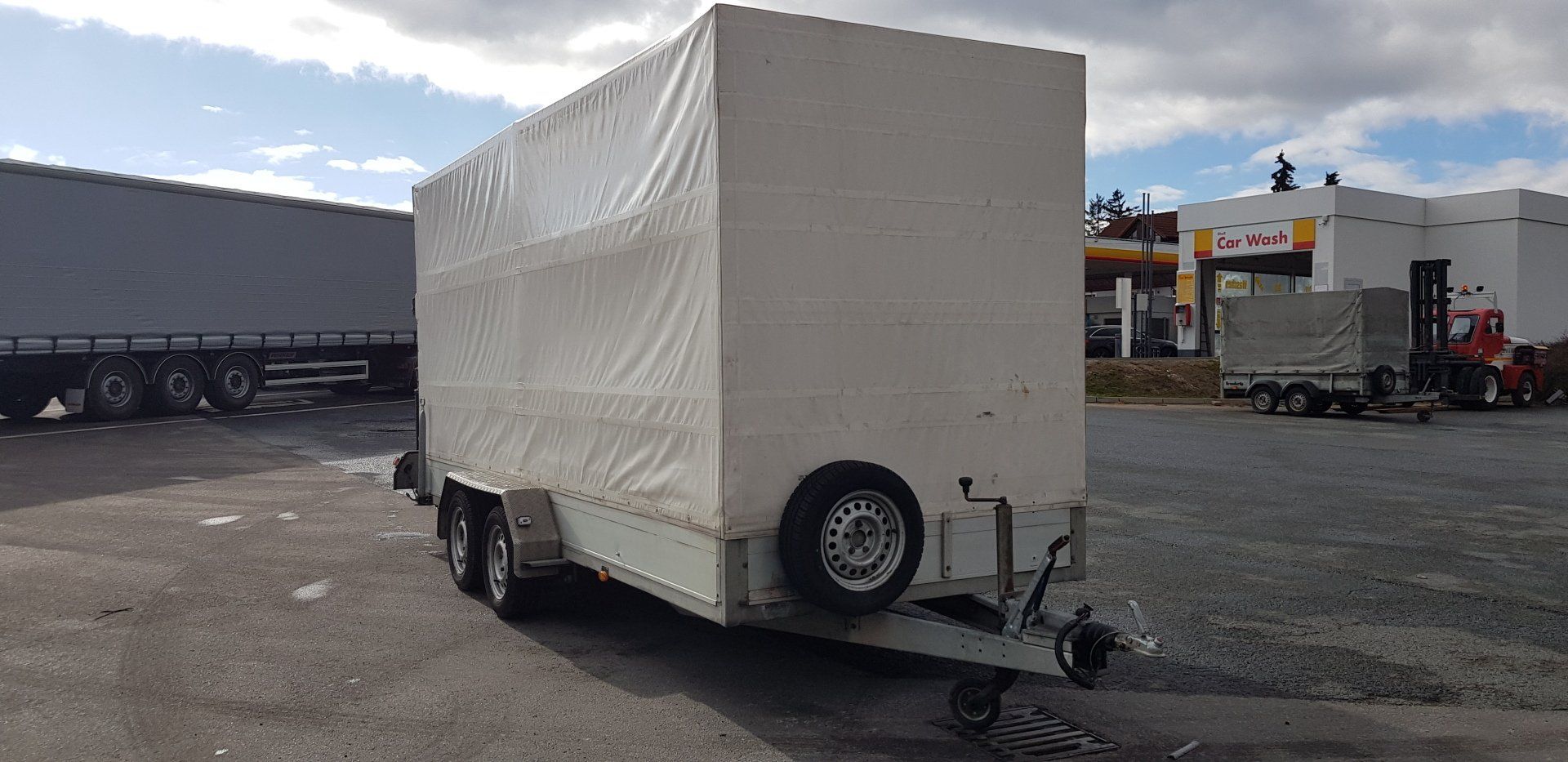 Geschlossener Anhänger mit weißer Abdeckung auf Asphalt geparkt; andere Fahrzeuge und Gebäude im Hintergrund.