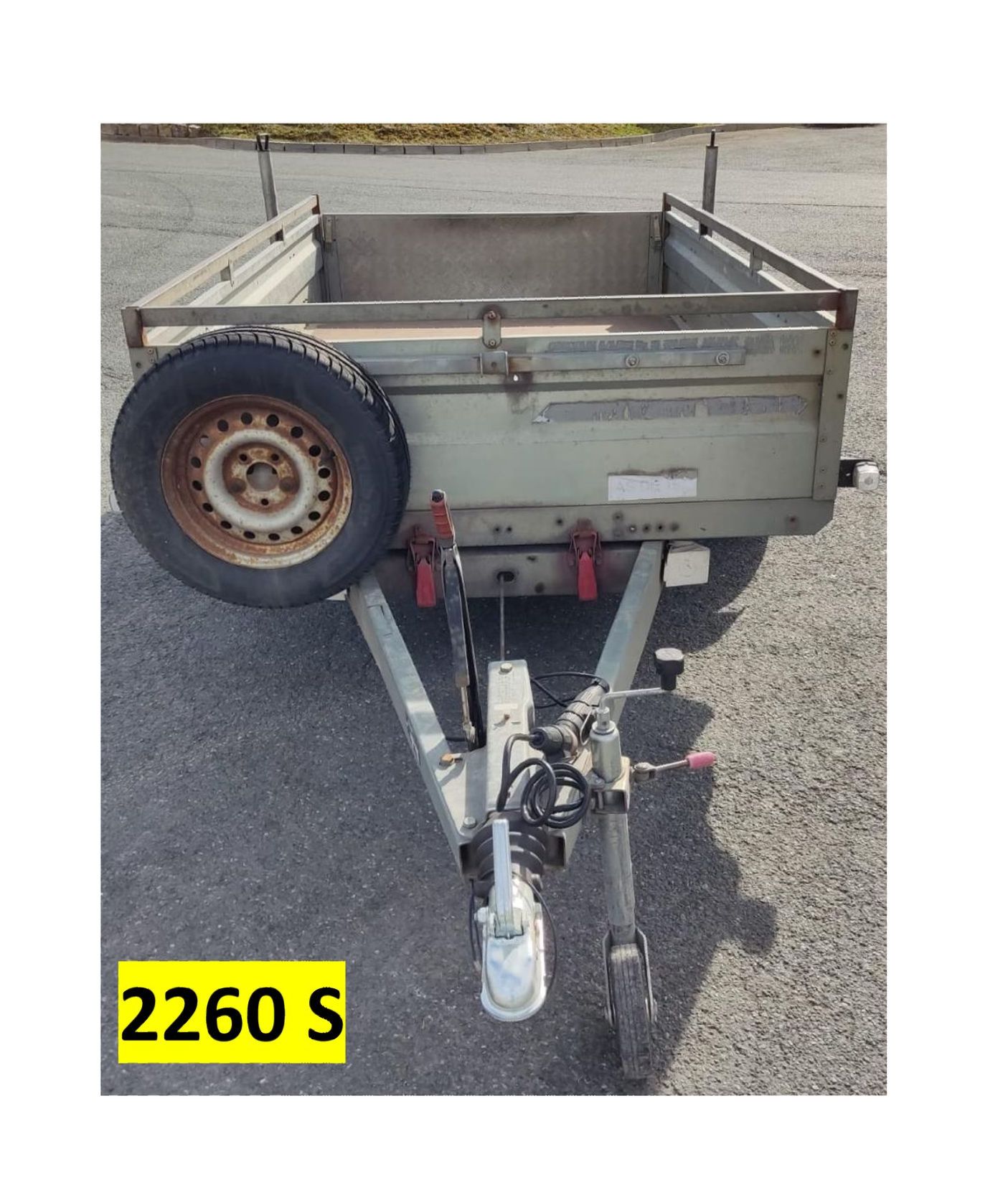 Grauer Nutzanhänger mit montiertem Ersatzreifen, geparkt auf Asphalt. Der Anhänger hat eine weiße Anhängerkupplung und ist mit „2260 S“ beschriftet.