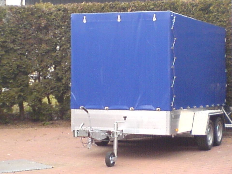 Geschlossener Anhänger mit blauer Abdeckung und silberner Metallbasis, an ein Fahrzeug in einer Gartenumgebung angehängt.