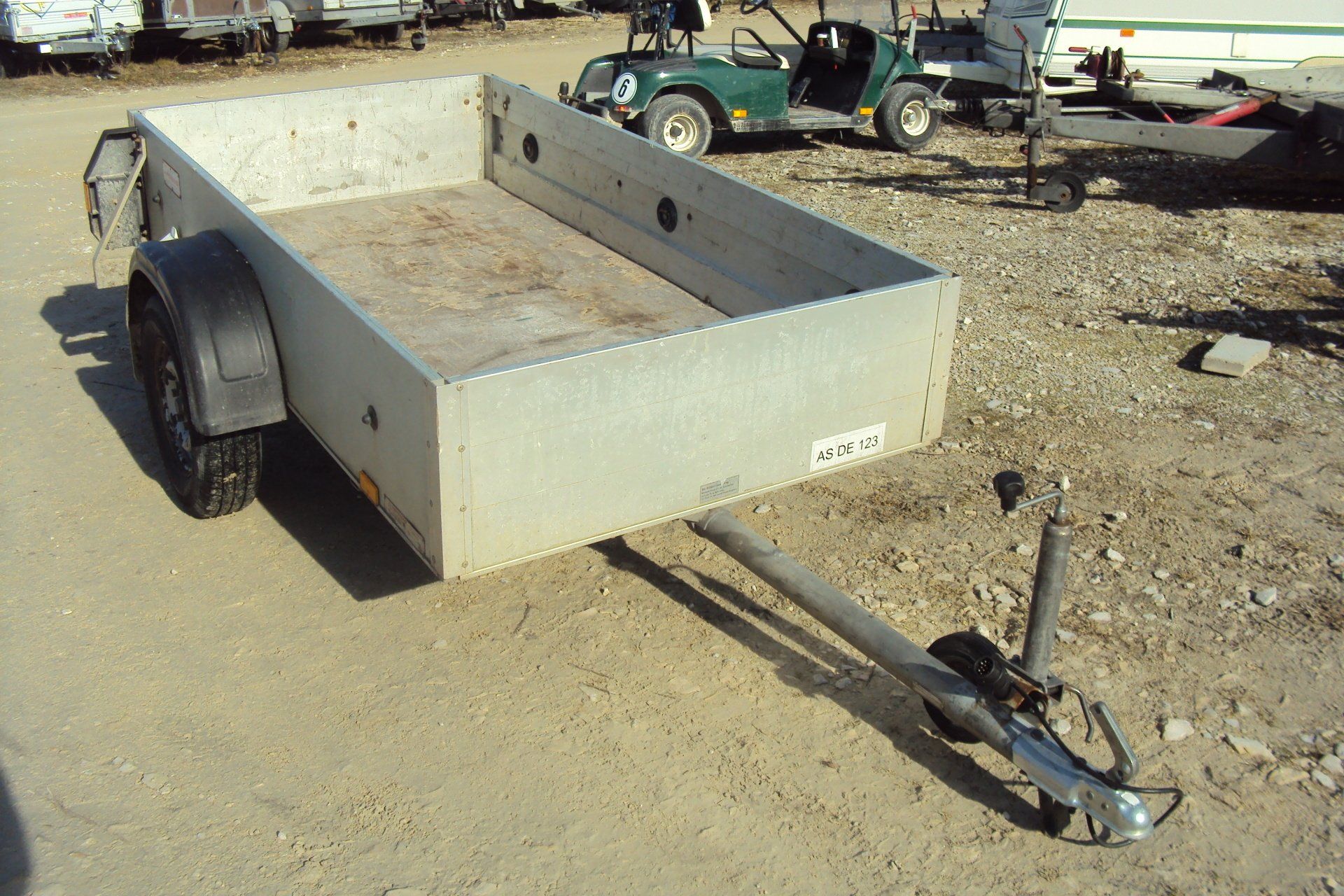 Silberner Nutzanhänger auf unbefestigter Fläche, im Hintergrund ein Golfwagen und andere Anhänger.