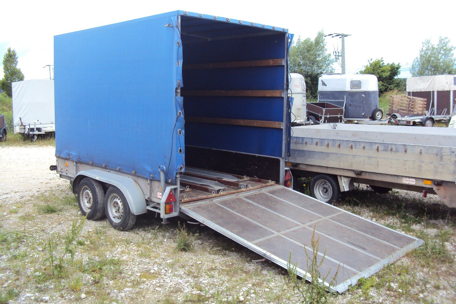 Blauer geschlossener Frachtanhänger mit Rampe, im Freien auf Schotter geparkt.