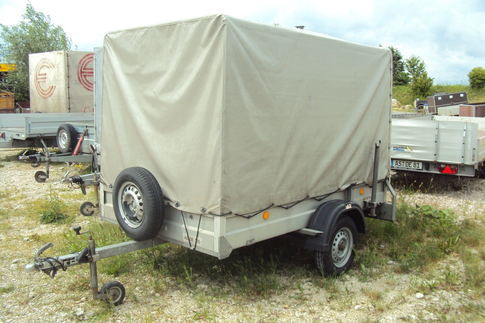 Geschlossener Nutzanhänger mit hellbrauner Abdeckung, Ersatzreifen und Anhängerkupplung, auf Gras geparkt.