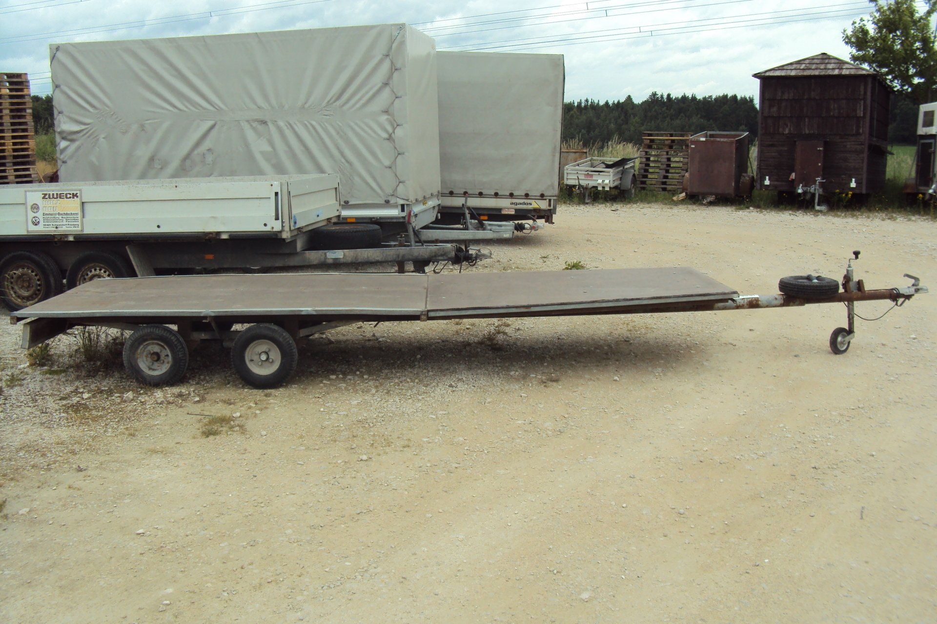 Ein Pritschenanhänger auf einem Schotterplatz, im Hintergrund sind Anhänger mit grauen Planen bedeckt.