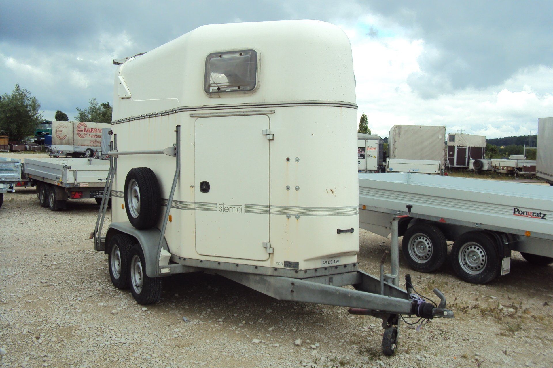 Weißer Pferdeanhänger mit angebrachtem Ersatzreifen, draußen mit anderen Anhängern geparkt.