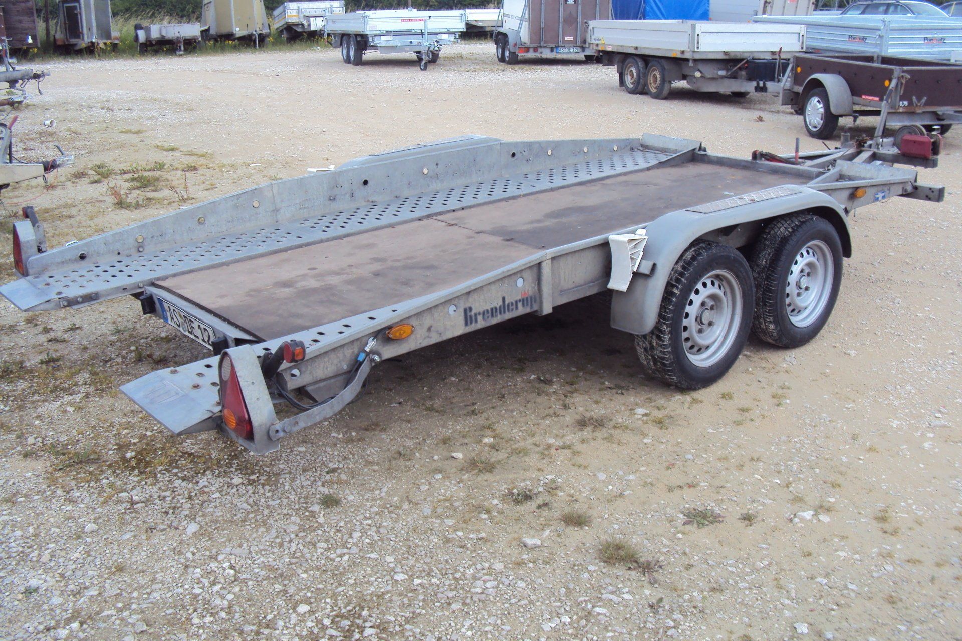 Autoanhänger mit Zwillingsrädern, Laderampe und Holzdeck, auf Schotter geparkt.