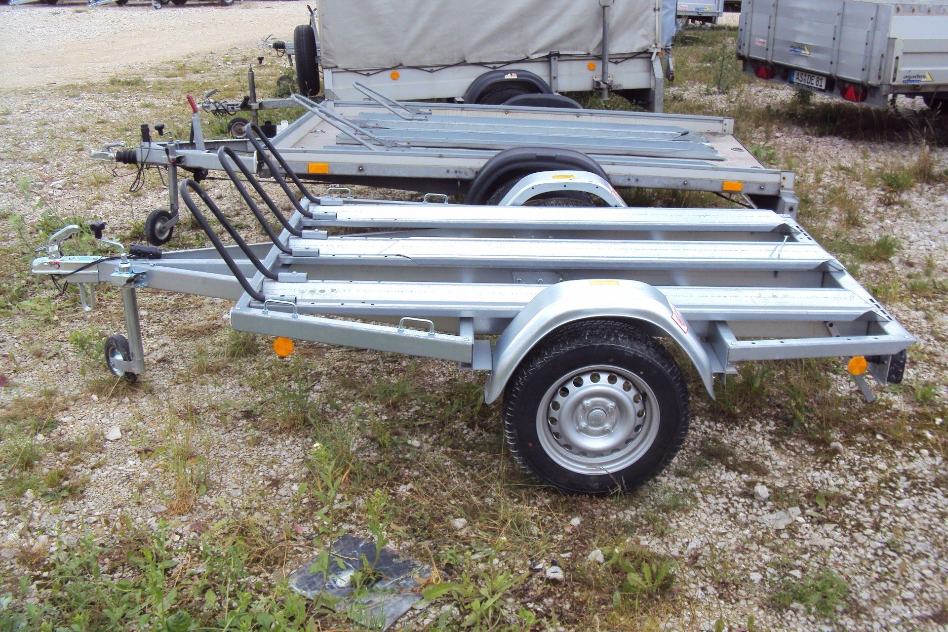 Ein Motorradanhänger auf einer Schotterfläche, mit schwarzem Reifen und Stahlrahmen.