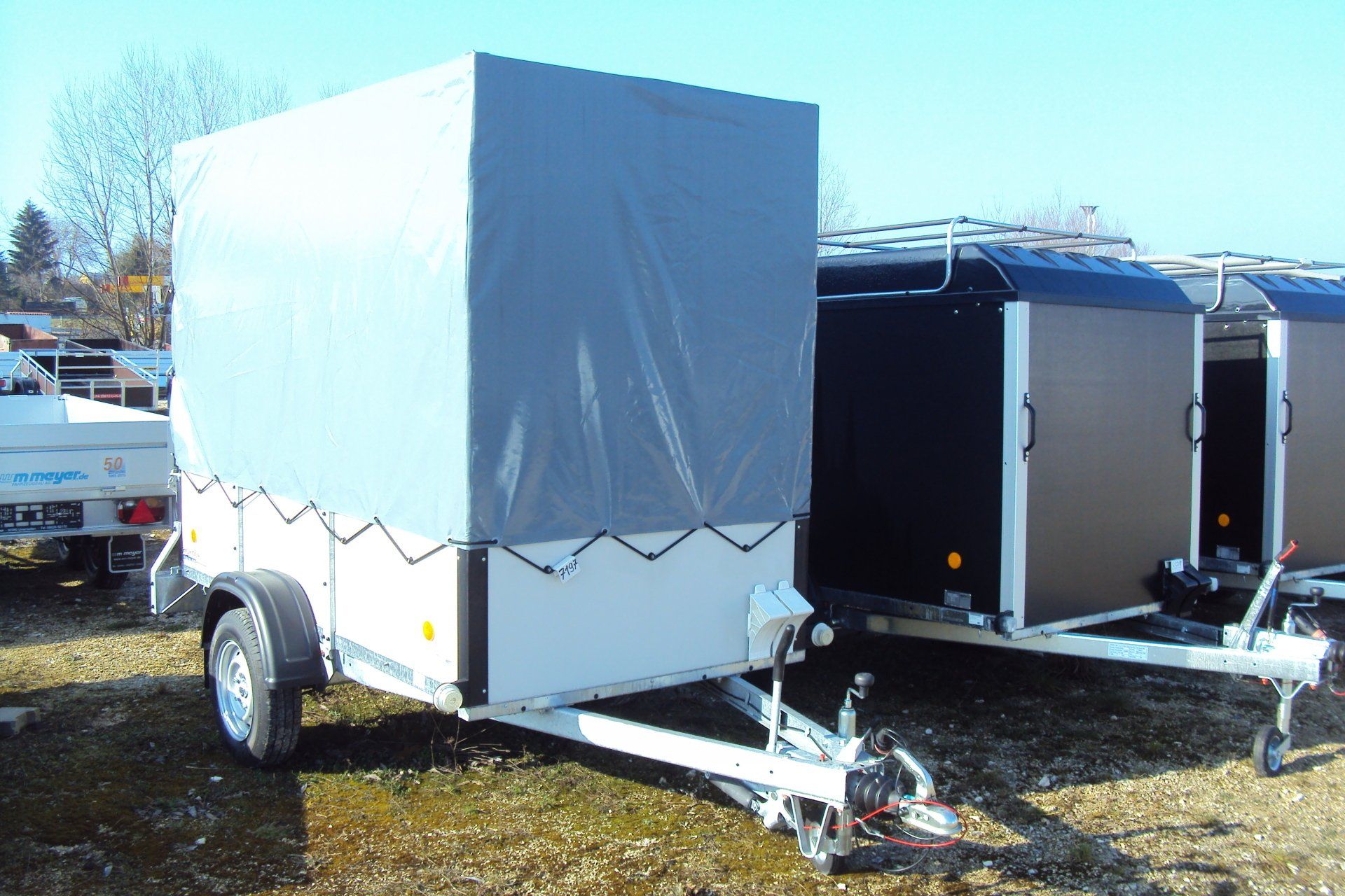 Weißer Nutzanhänger mit grauer Plane, der an einem sonnigen Tag draußen geparkt ist.