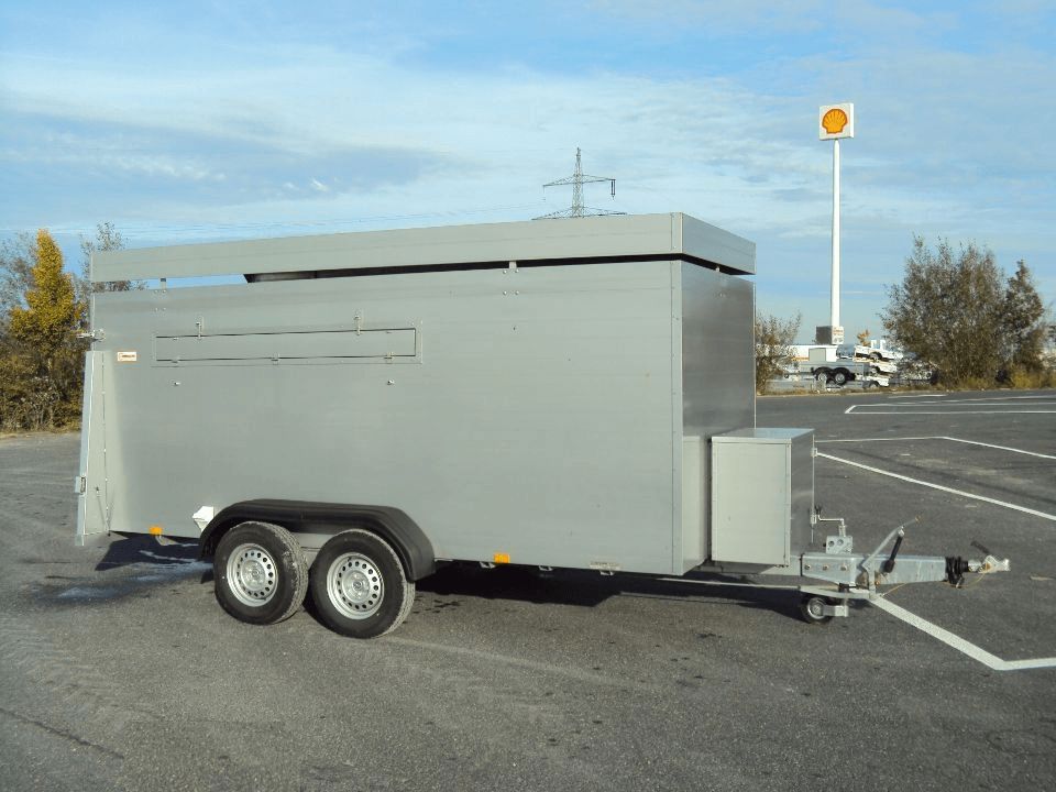Grauer geschlossener Anhänger mit Doppelrädern, im Freien geparkt, im Hintergrund eine Shell-Tankstelle.