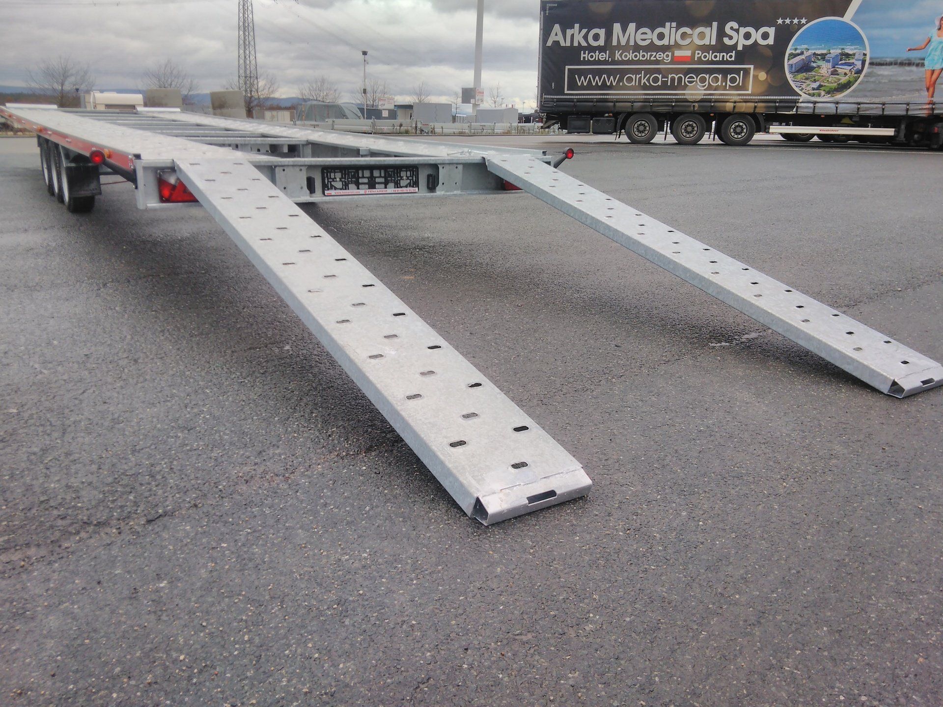 Ein Autotransportanhänger mit zwei Aluminiumrampen auf Asphalt. Im Hintergrund ist ein Sattelschlepper zu sehen.